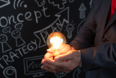 Man holding glowing lightbulb next to a blackboard with ideas on it
